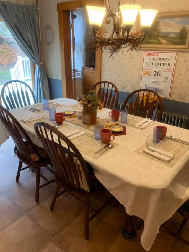 A dining room with a rectangular table covered with a white tablecloth set for six people. The table has red mugs, blue glasses, plates with butter, and silverware neatly arranged. A floral centerpiece is in the middle of the table. Wooden chairs surround the table. A window with blue curtains is on the left, and a calendar on the wall shows the date as Thursday, November 26. A light fixture with multiple bulbs hangs above the table.