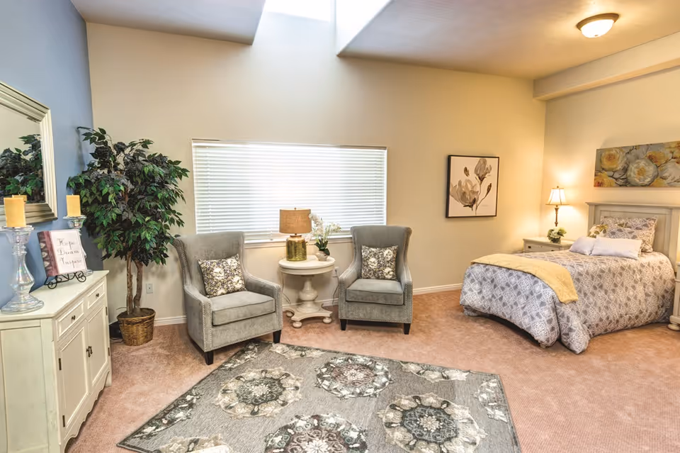 A cozy bedroom with a single bed covered in patterned bedding and a yellow throw blanket. Next to the bed is a nightstand with a lamp and a small plant. Two gray armchairs with floral cushions are positioned around a round white side table with a lamp and flowers. A large window with blinds is behind the chairs. A decorative rug with floral patterns covers part of the carpeted floor. A large mirror, candles, and a potted plant are on a white cabinet against the wall.