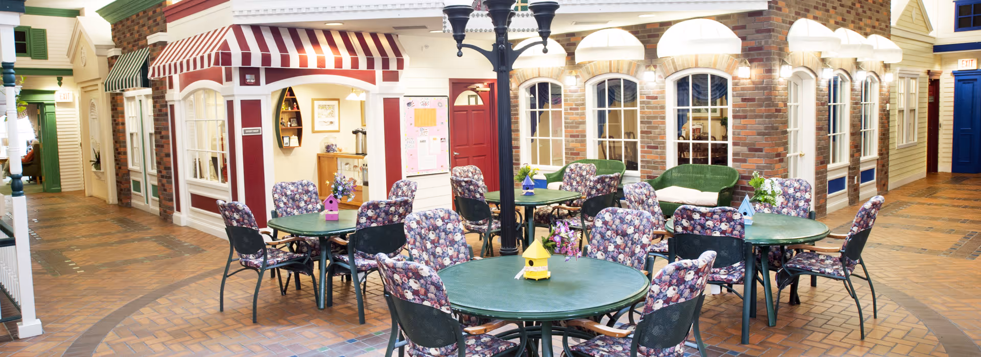 Indoor seating area with round tables and floral-patterned chairs arranged on a brick floor. The space resembles a small indoor village with storefront-style facades, including a red and white striped awning, windows, and doors. There are decorative birdhouses on the tables and a black streetlamp in the center. The setting is bright and inviting, designed to mimic an outdoor street scene inside a building.