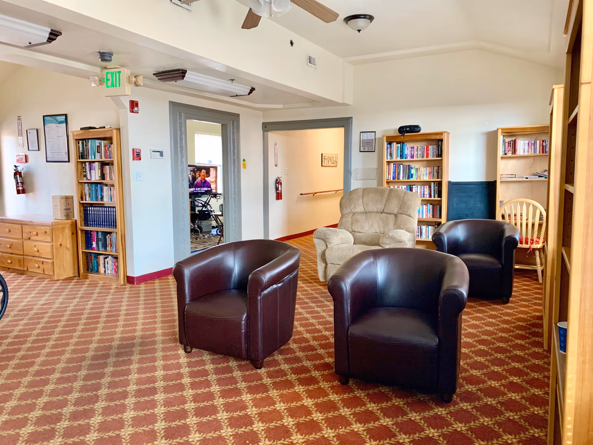 A cozy common area in an assisted living facility with three dark brown leather armchairs and one beige recliner arranged on a patterned carpet. There are wooden bookshelves filled with books along the walls, a wooden dresser, and a wooden chair with a cushion. The room has white walls and ceiling fans, with an exit sign and fire extinguisher visible near the doorways.