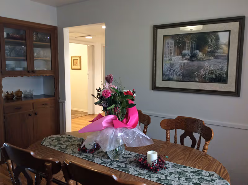 A dining room with a wooden table and six matching chairs. On the table is a floral table runner, a vase with a bouquet of flowers wrapped in pink and clear plastic, and a candle centerpiece with red berries. To the left is a wooden cabinet with glass doors displaying dishes. A framed painting of an outdoor garden scene hangs on the wall. The room has light-colored walls and a doorway leading to a hallway with wooden flooring.