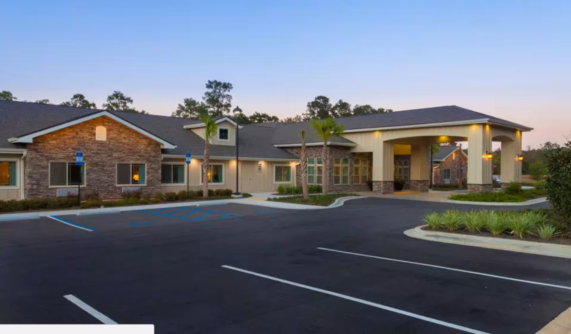 Exterior view of a single-story senior living facility building with stone and beige siding, a covered entrance, palm trees, and a parking lot with marked handicap spaces during dusk.