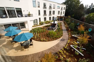Outdoor courtyard area at Aegis Living West Seattle featuring a garden with various plants, a wooden pathway, patio tables with blue umbrellas, and a decorative boat centerpiece. The multi-story building with balconies overlooks the courtyard.