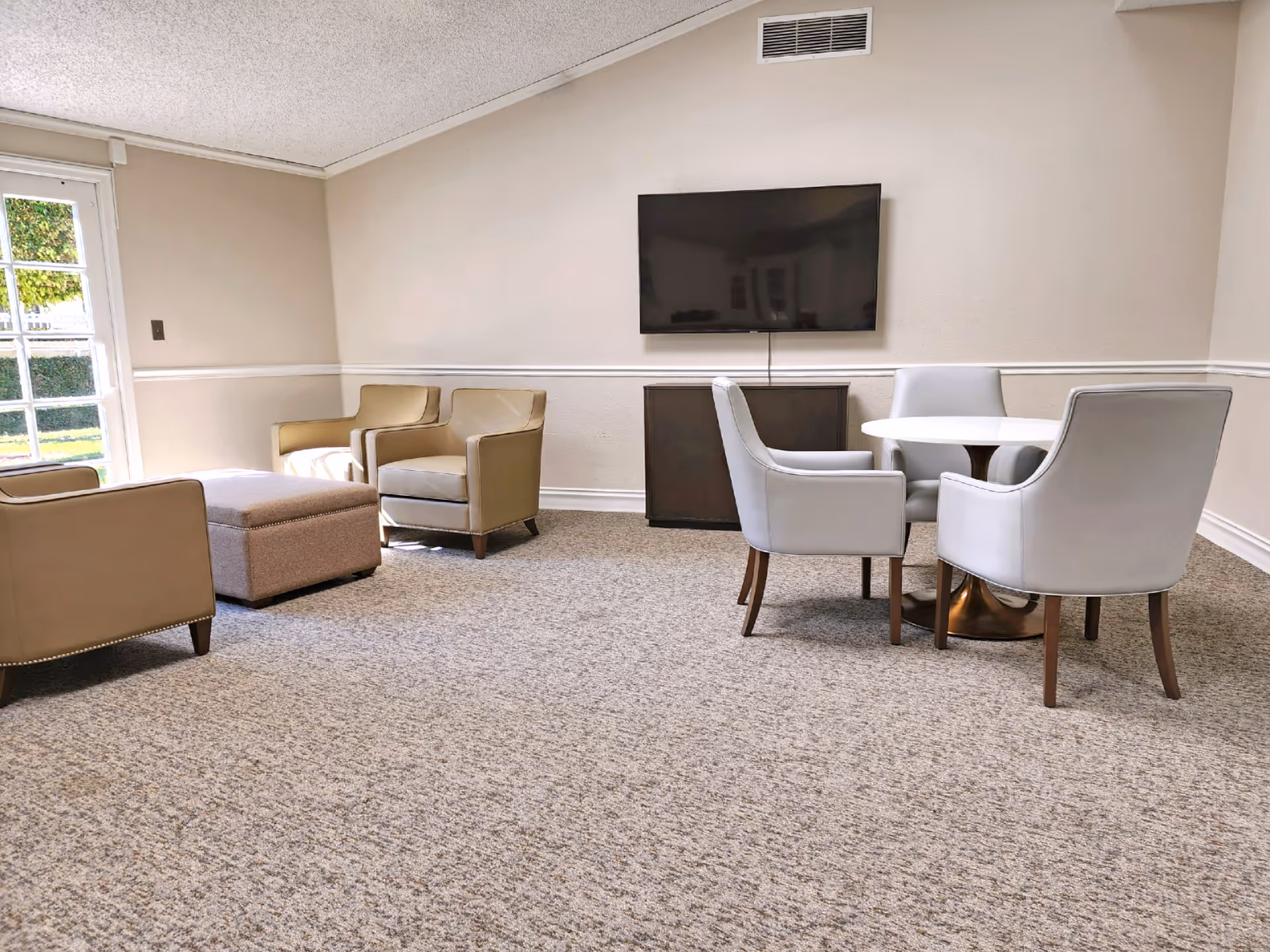 A cozy sitting area in a memory care facility with beige armchairs, a matching ottoman, a round white table surrounded by four white chairs, a wall-mounted flat-screen TV, and a door with glass panes letting in natural light.
