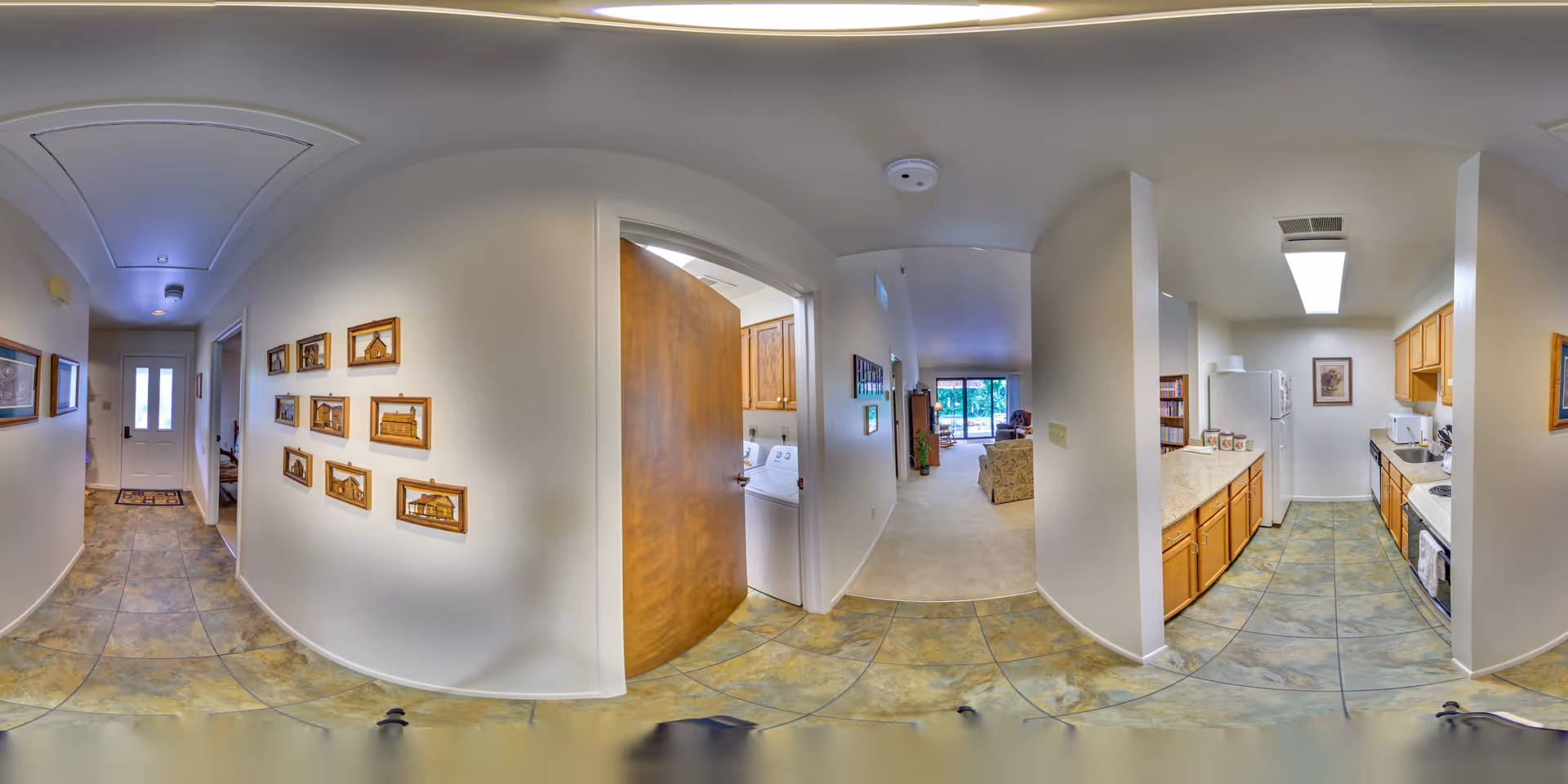 A wide-angle view of an interior hallway in a senior living facility showing a tiled floor leading to a white front door. On the left wall, there are framed pictures. To the right, an open door reveals a laundry area with a washer and dryer. Further right is a kitchen with wooden cabinets, a refrigerator, microwave, and stove. Beyond the hallway is a carpeted living room area with sofas and large windows letting in natural light.