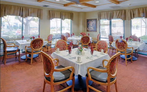 A dining room with multiple tables covered in white tablecloths, each set with cups, utensils, and napkins. The chairs have wooden frames with red patterned upholstery. Large windows with beige valances let in natural light, and a framed painting hangs on the wall.