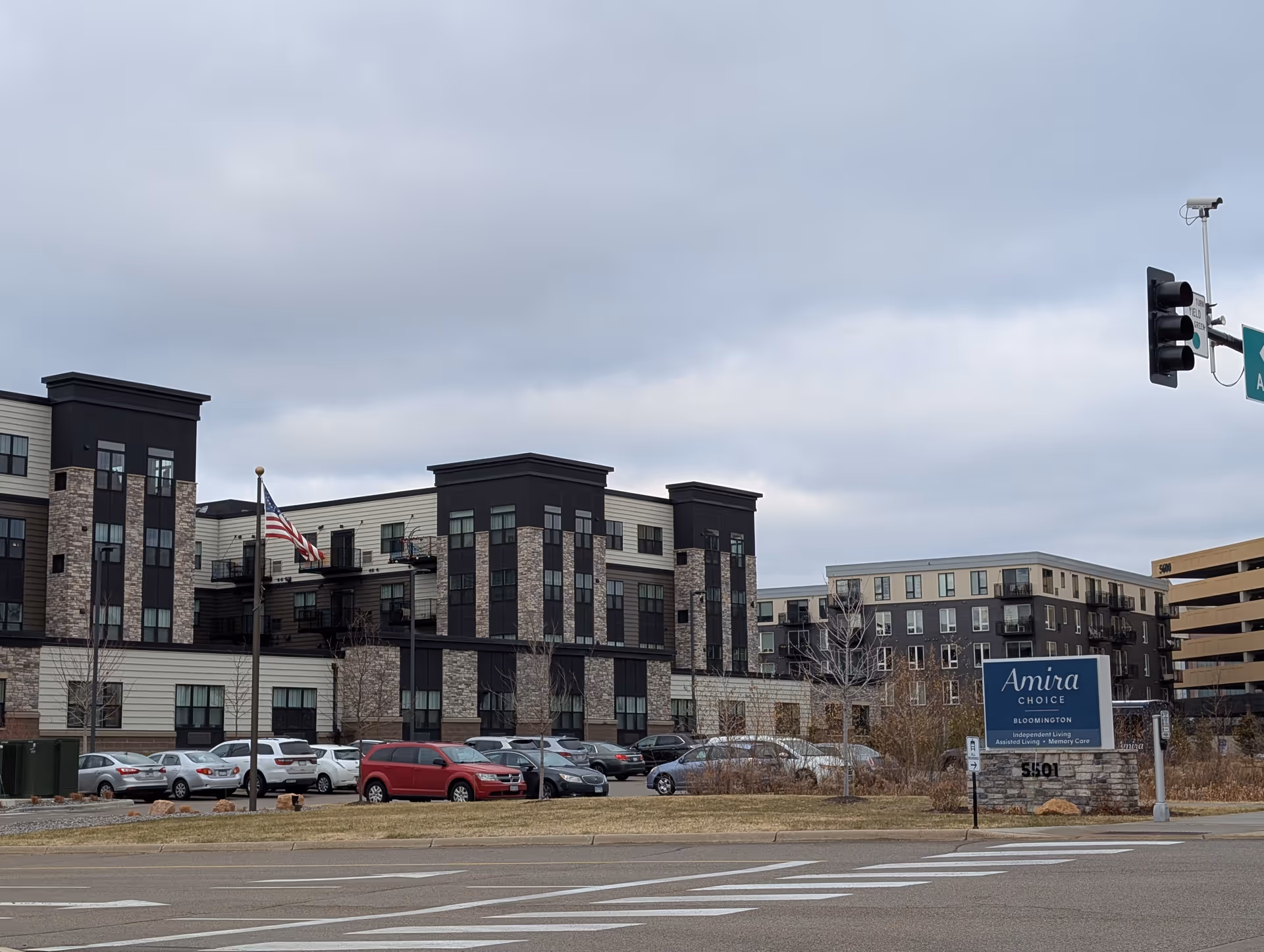 Exterior view of Amira Choice Bloomington senior living facility with a parking lot in front, an American flag on a flagpole, and a cloudy sky overhead.