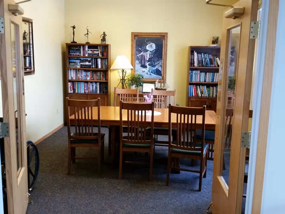 A cozy room with a wooden table surrounded by six wooden chairs. There are two bookshelves filled with books against the back wall, a floor lamp, and a framed painting of a person reading. The room has beige walls and carpeted floor, viewed through open double wooden doors.