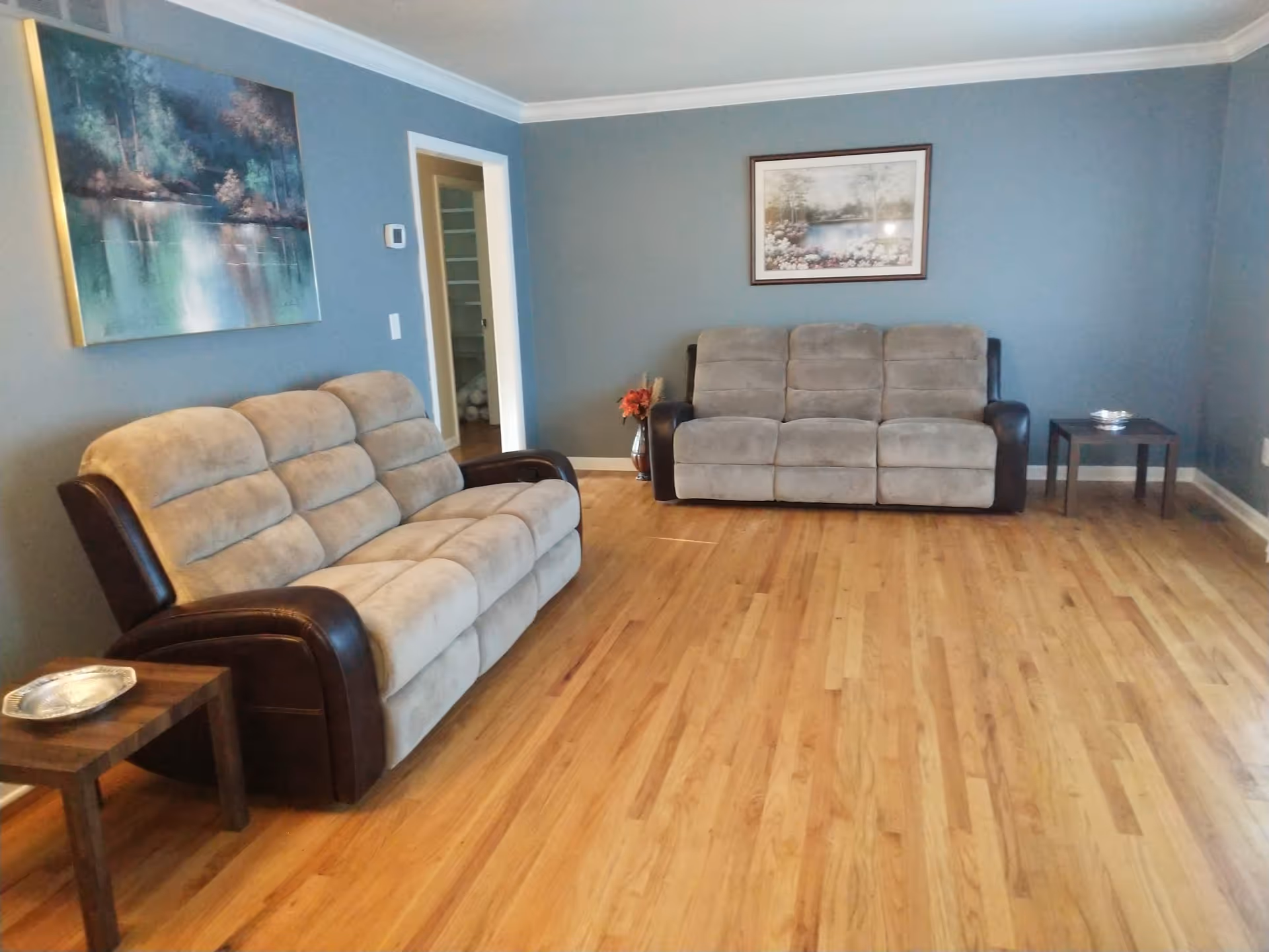 Spacious living room with two beige-and-brown reclining sofas, hardwood floors, side tables, and framed wall art on blue-gray walls.