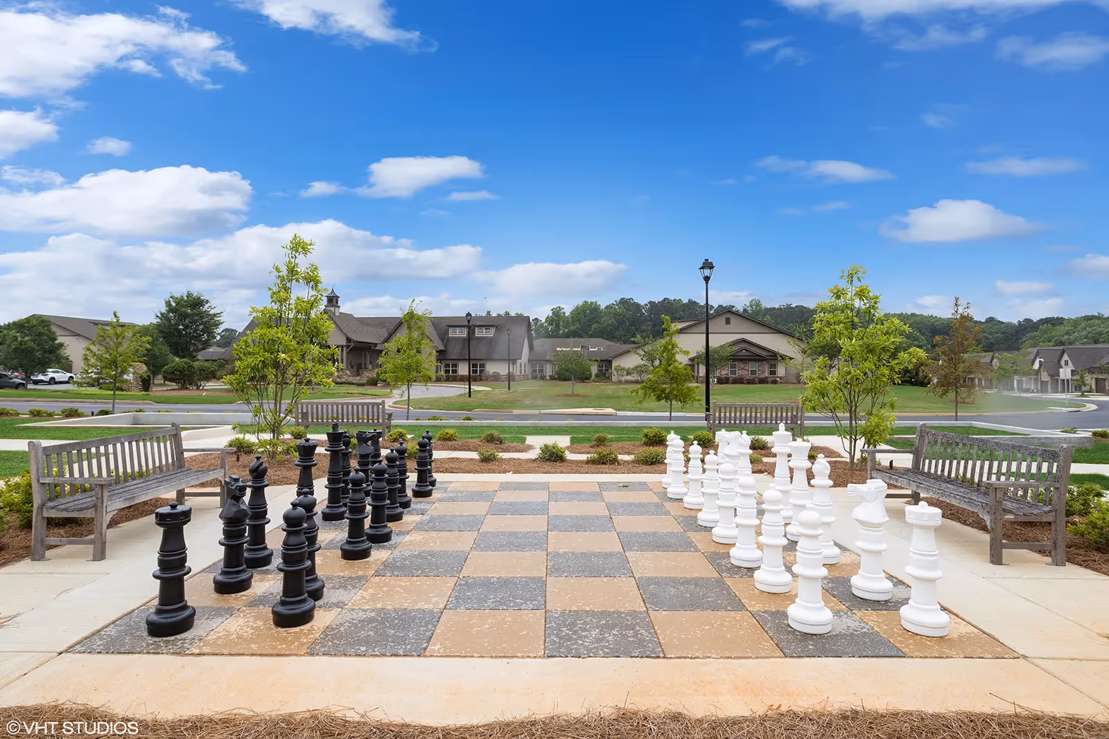 Outdoor area with a large chessboard featuring oversized black and white chess pieces set up for a game. Surrounding the chessboard are wooden benches and small trees, with a view of residential buildings and a clear blue sky with scattered clouds in the background.