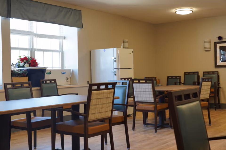 A dining area with multiple wooden tables and chairs with padded seats and backs. There is a large window with a valance letting in natural light, and a white refrigerator is visible against the beige wall. The floor is wooden, and there is a framed picture on the wall along with ceiling and wall lights.