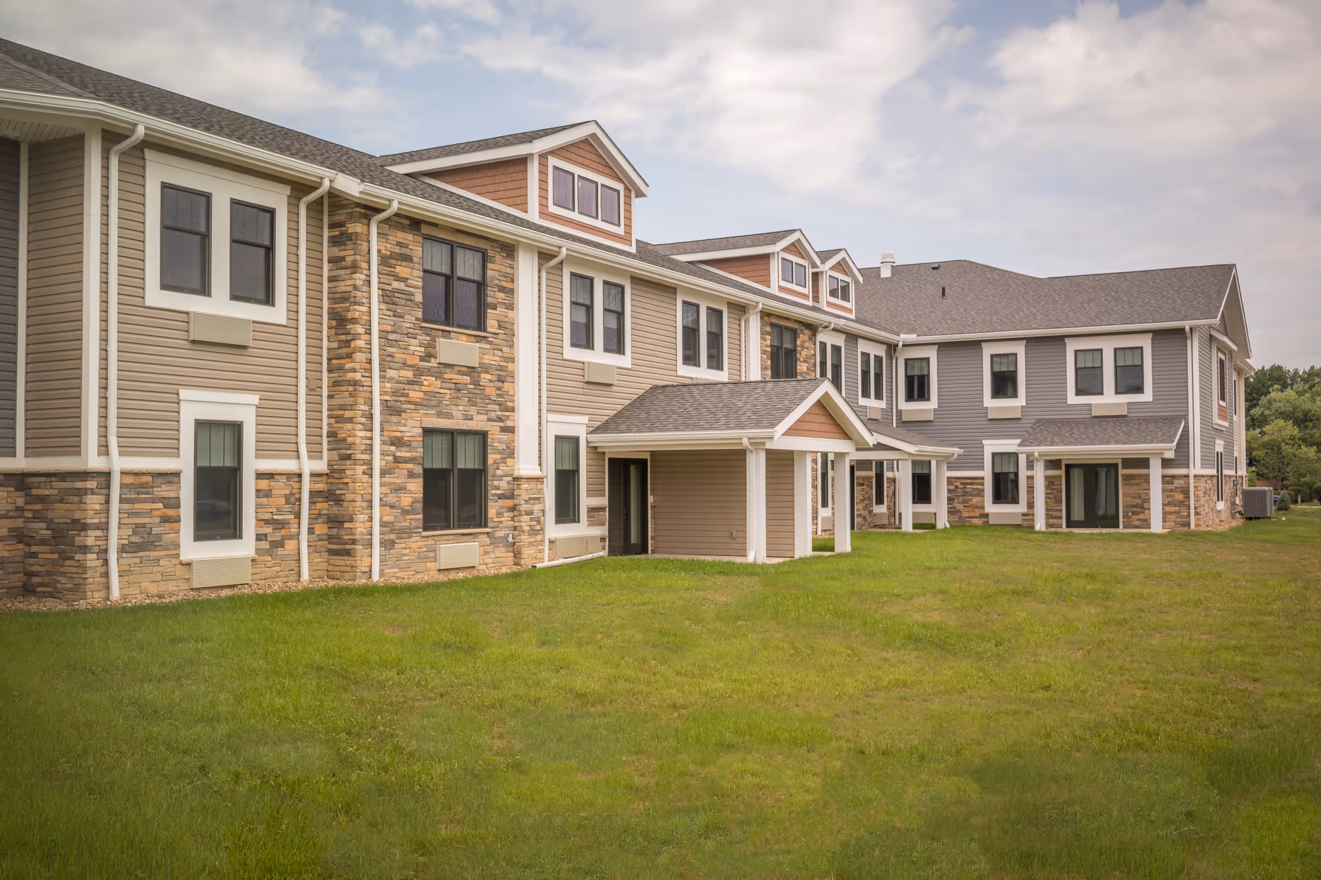 Two-story senior living building with stone and siding exterior, multiple windows, and a grassy lawn.