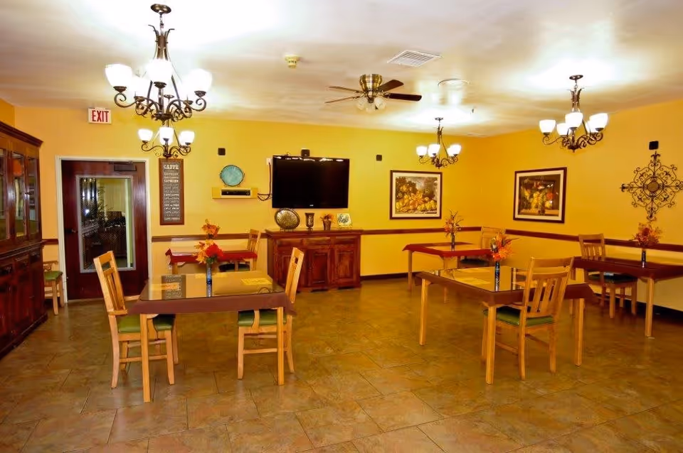 A dining room with several wooden tables and chairs arranged neatly. Each table has a small vase with orange flowers. The walls are painted yellow and decorated with framed artwork and a decorative metal wall piece. There are multiple chandeliers and ceiling fans providing lighting. A flat-screen TV is mounted on the wall above a wooden cabinet. An exit door is visible on the left side of the room.