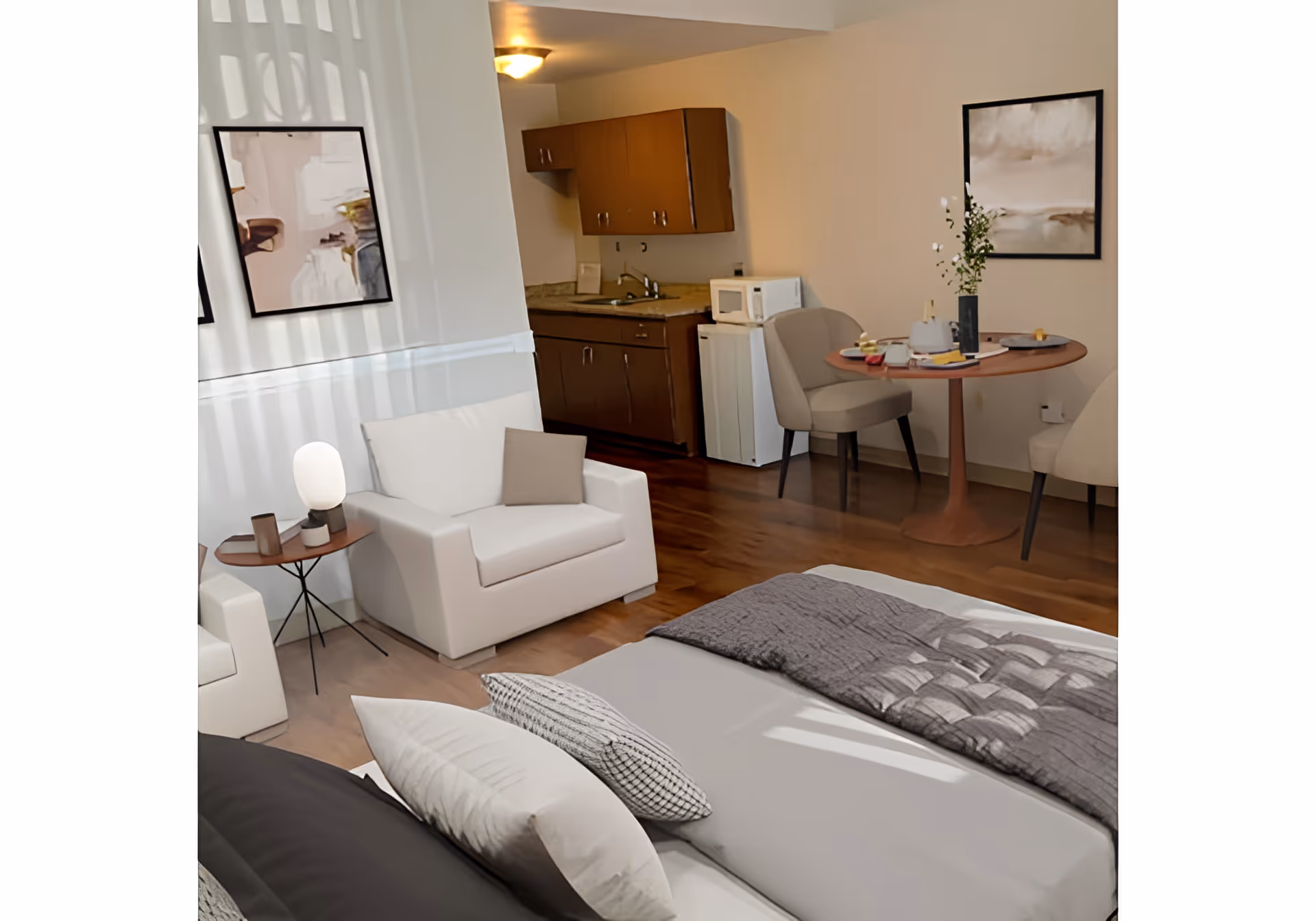 A cozy studio apartment featuring a bed with pillows and a gray quilt, a white armchair with a small side table and lamp, a round dining table with two chairs, a small kitchenette with wooden cabinets, a sink, a microwave, and a mini refrigerator. The walls are decorated with framed abstract artwork.