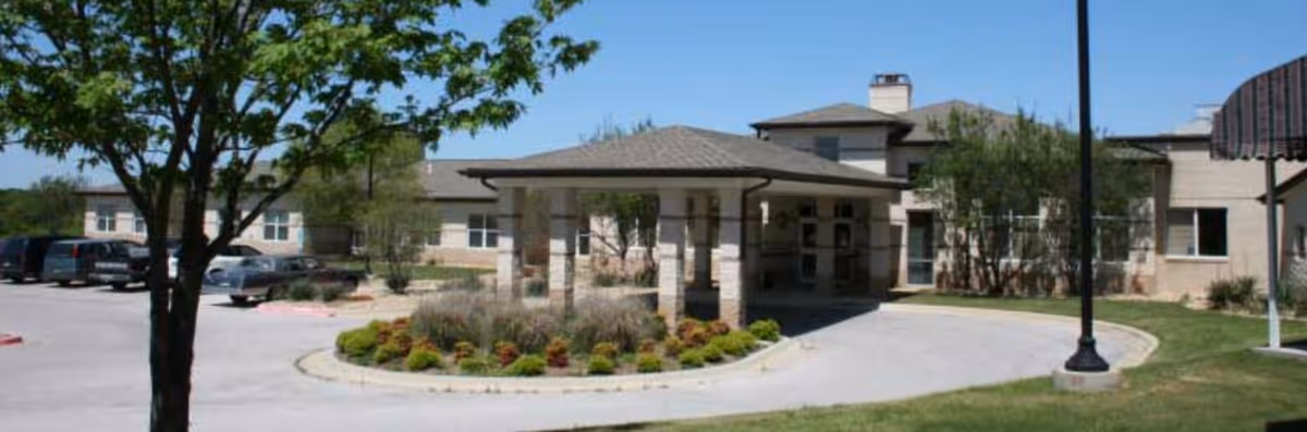 Exterior view of Coryell Memorial Healthcare System Residential Care building with a covered entrance, landscaped garden area, parking lot with several cars, and a tree in the foreground under a clear blue sky.