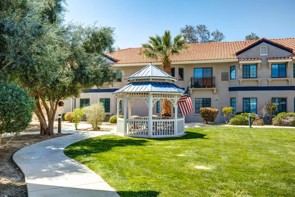 Outdoor area of The Havens at Antelope Valley featuring a white gazebo with an American flag, surrounded by green grass, trees, and shrubs, with a two-story building with a tiled roof in the background.