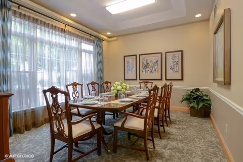 A formal dining room with a long wooden table set for eight people. The table has plates, glasses, and a centerpiece of white flowers. There are eight wooden chairs with cushioned seats around the table. The room has large windows with sheer curtains allowing natural light to enter. The walls are decorated with three framed floral paintings and a potted plant is placed in the corner.