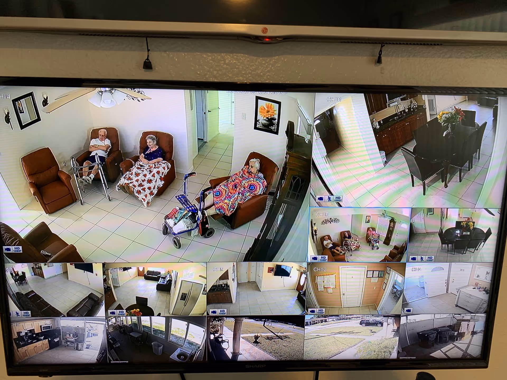 Security camera monitor showing multiple views of a senior living facility. The main view shows three elderly individuals seated in recliners in a tiled common area, two covered with blankets and one with a walker nearby. Other smaller views include a dining area with a table and chairs, hallways, an office area, and outdoor spaces with grass and trash bins.