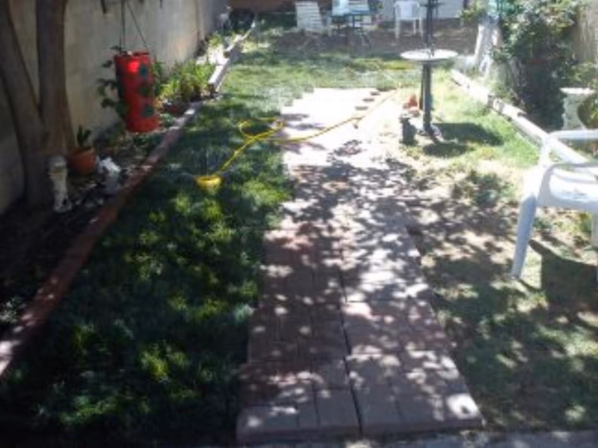 A shaded outdoor garden area with a brick pathway running through the middle, surrounded by grass and plants. There are white plastic chairs and a small round table in the background, along with a red hanging planter on the left side.