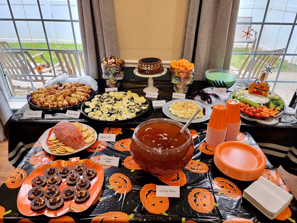 A Halloween-themed buffet table set up indoors near large windows with a view of outdoor chairs and grass. The table is covered with a black tablecloth decorated with orange jack-o'-lantern faces. Various Halloween-themed foods are displayed, including mummy dogs, deviled eggs labeled as spider eggs, a brain-shaped meatloaf called 'Horrors Meat' with crackers, chocolate cupcakes with orange sprinkles called 'Witches Hats', a bowl of punch labeled 'Witches Brew', a chocolate cake on a stand, bowls of snacks, and a vegetable platter with a carved pumpkin decoration. Orange plates, cups, and napkins are stacked on the table.