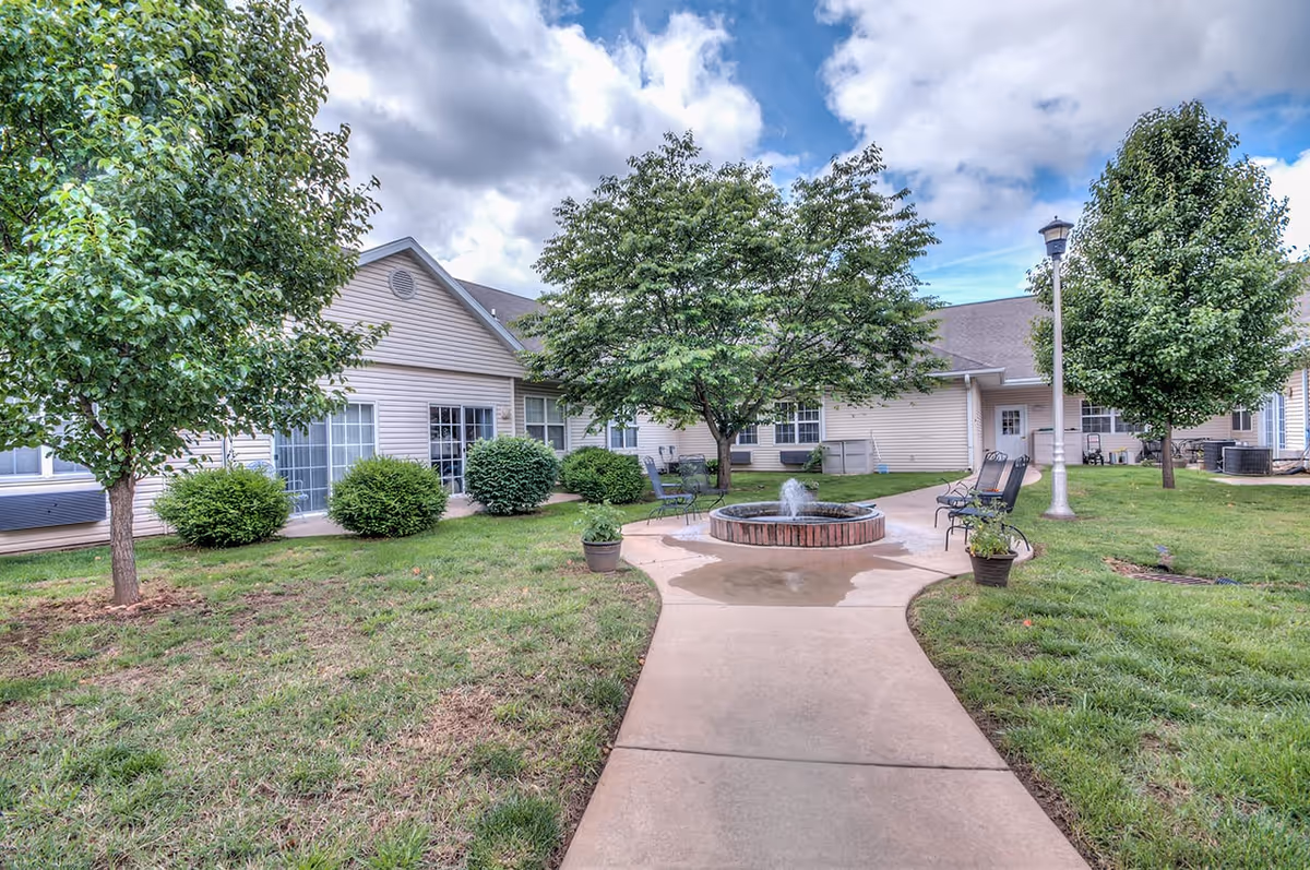 Outdoor courtyard area at The Bungalows at Chesterfield Village featuring a concrete walkway leading to a circular brick water fountain surrounded by green grass, trees, shrubs, and outdoor seating benches under a partly cloudy sky.