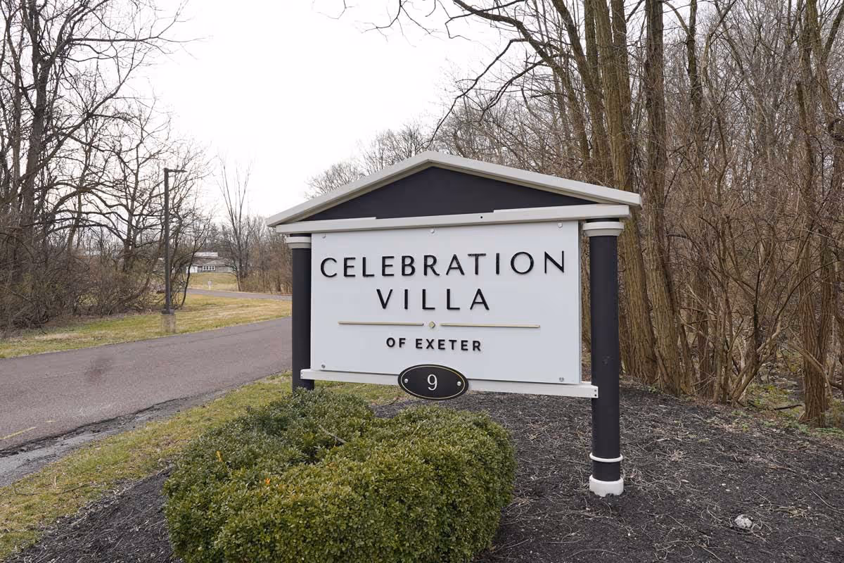 Outdoor view of a sign for Celebration Villa of Exeter, positioned next to a paved road with leafless trees and bushes in the background.