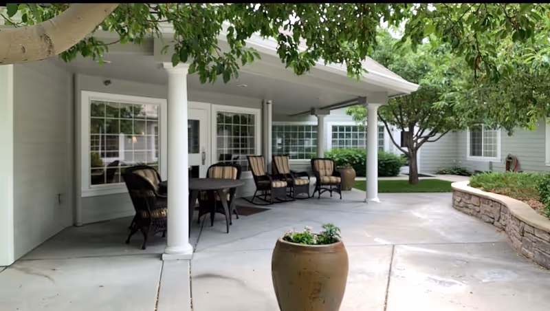 Covered outdoor patio area with several cushioned chairs and a round table. The patio is supported by white columns and overlooks a garden area with trees and shrubs. There is a large planter with flowers in the foreground and a stone retaining wall on the right side.