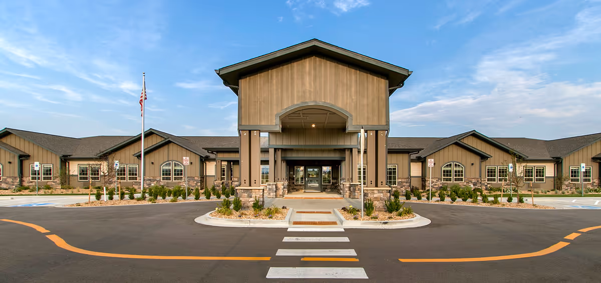 Front entrance of a single-story senior living facility with a covered porte-cochere, landscaped islands, and an American flag under a blue sky.