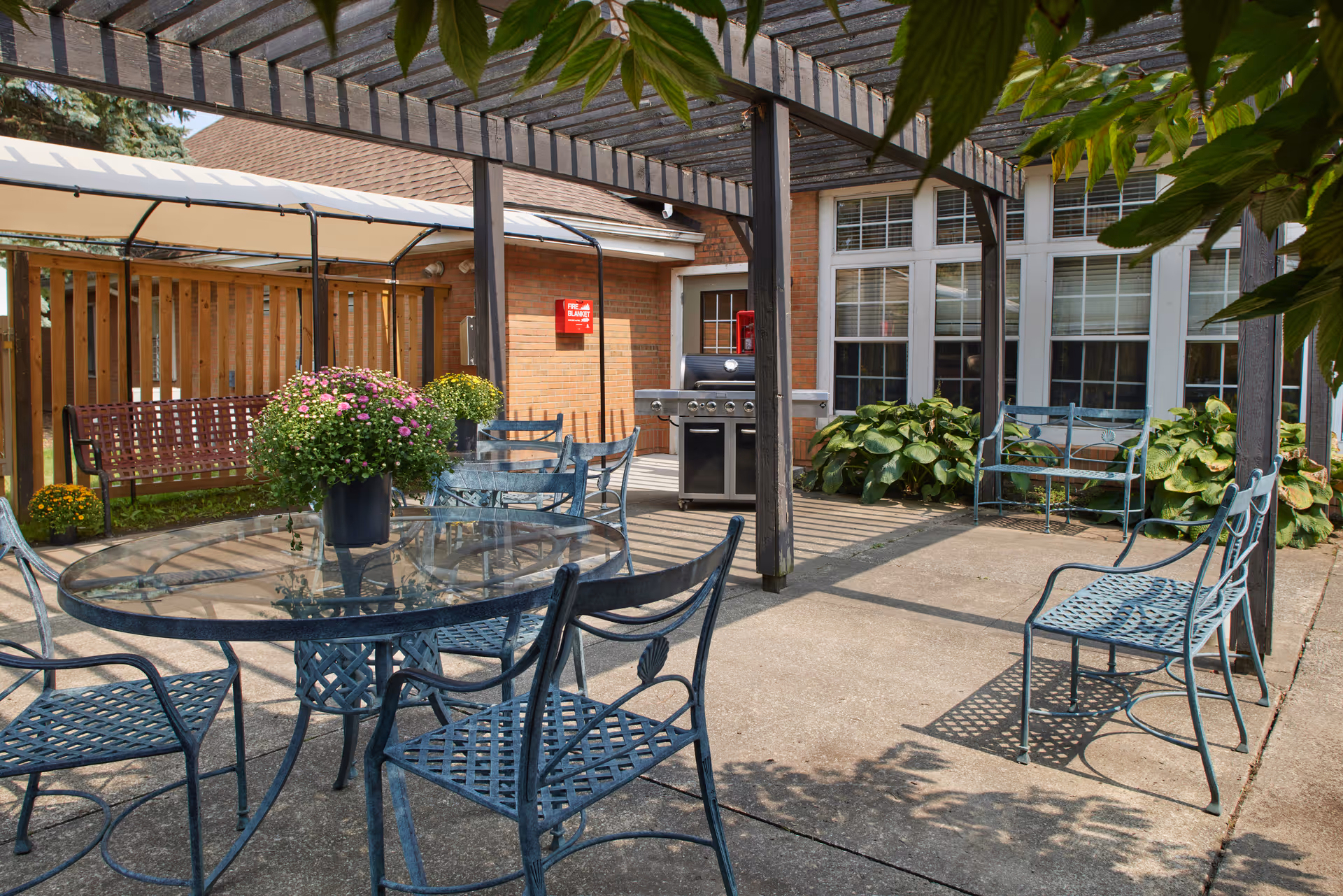 Outdoor patio area with a glass-top round table surrounded by metal chairs under a wooden pergola. There are potted flowers on the table, a bench swing with a canopy, a barbecue grill, and green plants near the building with large windows.
