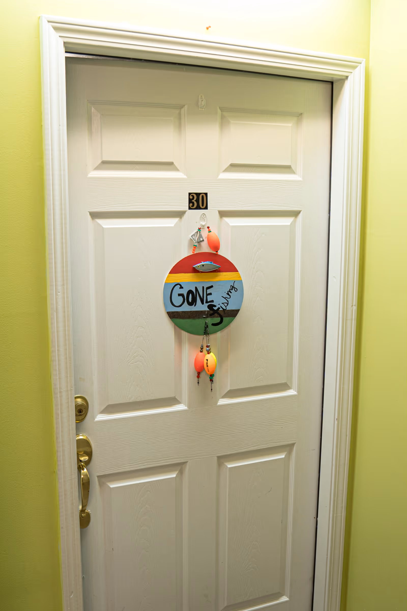 A white door with the number 30 on it, decorated with a colorful round sign that says 'Gone Fishing' and has fishing bobbers hanging from it. The door is set in a light green wall.