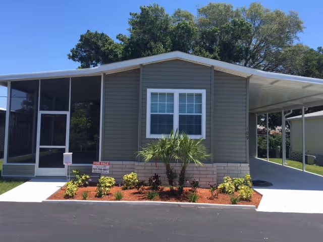 Single-story manufactured home with gray siding and white trim, featuring a screened porch on the left and a covered carport on the right. There is a small landscaped area with plants and a palm tree in front of the house. A 'For Sale' sign is visible near the porch. Trees are in the background under a clear blue sky.
