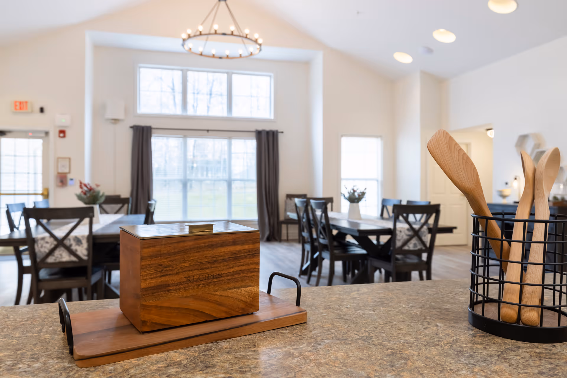 A bright dining room with multiple wooden tables and chairs, large windows letting in natural light, a chandelier hanging from the ceiling, and a countertop in the foreground holding a wooden recipe box and a black wire container with wooden cooking utensils.