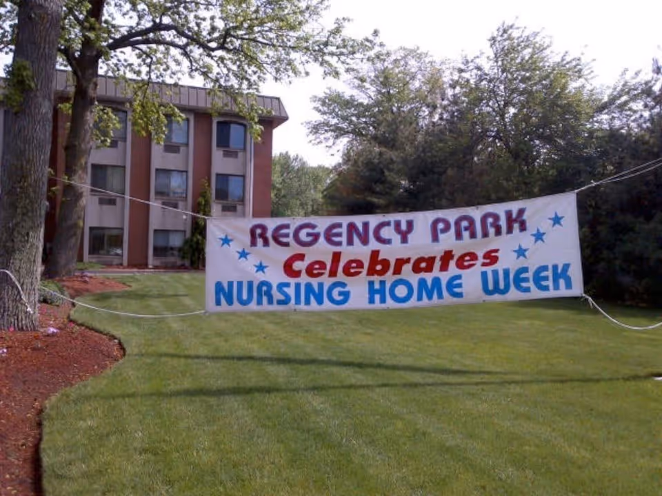 A large white banner with blue and red text reading 'REGENCY PARK Celebrates NURSING HOME WEEK' is displayed outdoors on a grassy lawn with trees and a multi-story building in the background.