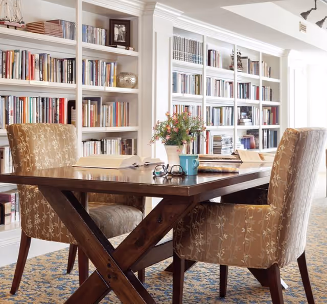 A cozy reading or study area with a wooden table surrounded by upholstered chairs with floral patterns. The table has an open book, a pair of glasses, a blue mug, and a vase with flowers. Behind the table is a large built-in bookshelf filled with books and decorative items.