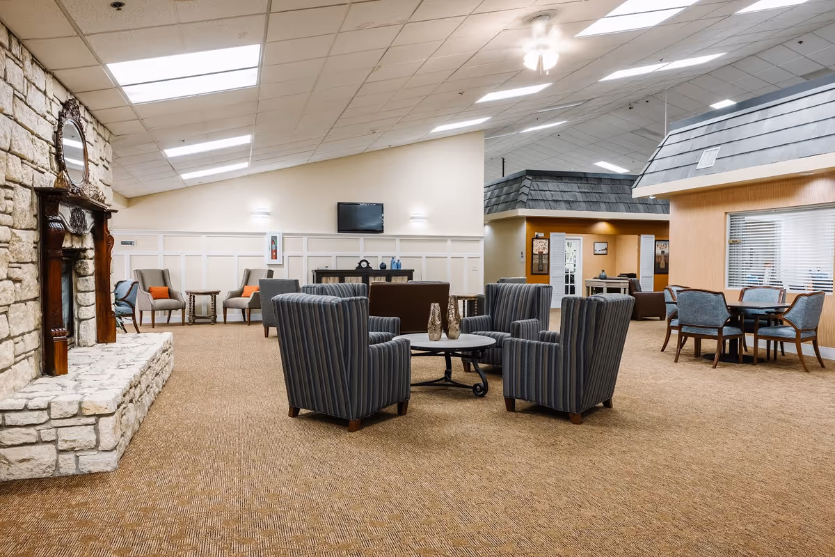 A spacious senior living common area with a stone fireplace on the left, four striped armchairs arranged around a round coffee table in the center, and additional seating areas with chairs and tables in the background. The room has a beige carpet, white paneled walls, and a ceiling with recessed lighting and a ceiling fan.