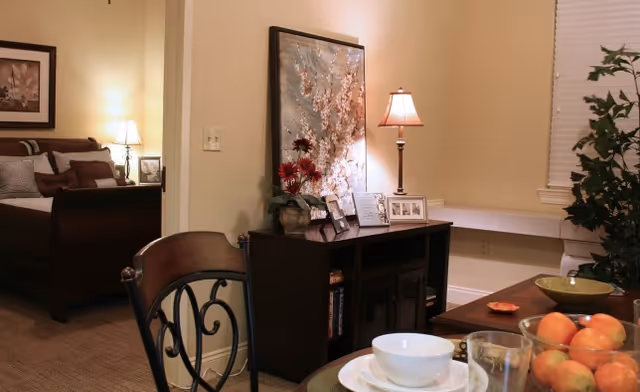 View of a cozy living and dining area in a senior living facility. The foreground shows a dining table set with bowls, glasses, and a bowl of fruit. Behind the table is a dark wooden cabinet with framed photos, a lamp, and decorative flowers. To the left, a bedroom with a bed, pillows, and a lamp is partially visible through an open doorway. The walls are painted a warm beige color, and there is a window with blinds and a green plant near the corner.