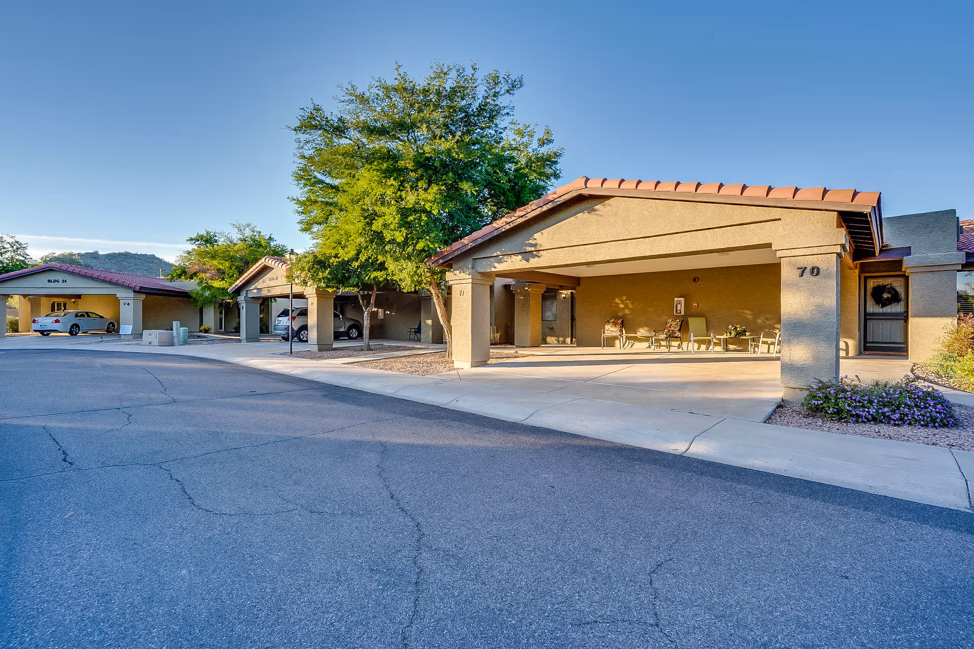 Exterior view of a senior living facility with covered parking spaces and a seating area under a roofed structure. Several cars are parked, and there are trees and shrubs around the buildings. The sky is clear and blue.