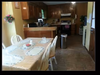 Dining table with placemats in the foreground and a kitchen with wooden cabinets, countertop, and stove in the background.