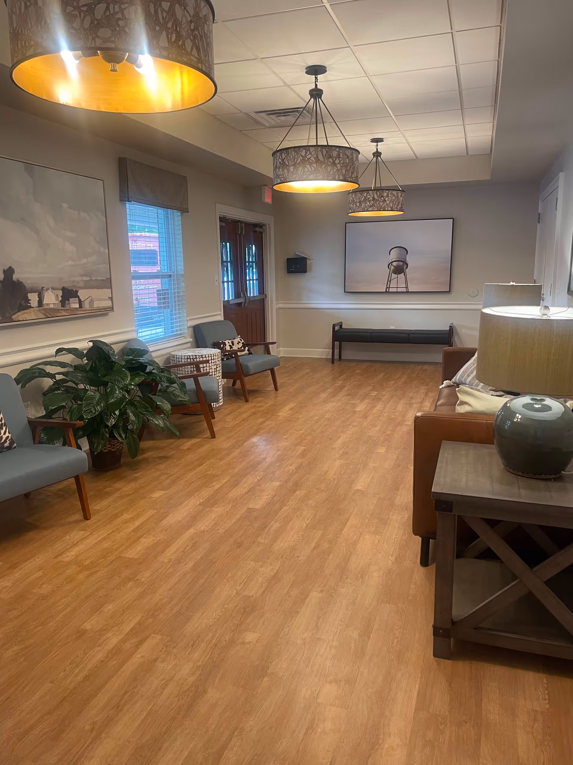 A well-lit waiting or sitting area with wooden flooring, featuring several chairs with cushions, a brown leather sofa, a side table with a lamp, two large paintings on the walls, three hanging ceiling lights, a potted plant, and a window with blinds.
