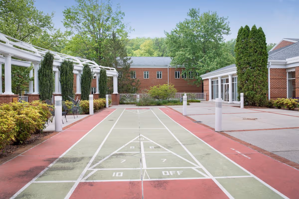 Outdoor shuffleboard court with red and green painted surface, surrounded by landscaped bushes, tall trees, and white columns with benches along the left side. Brick buildings and a clear blue sky are visible in the background.
