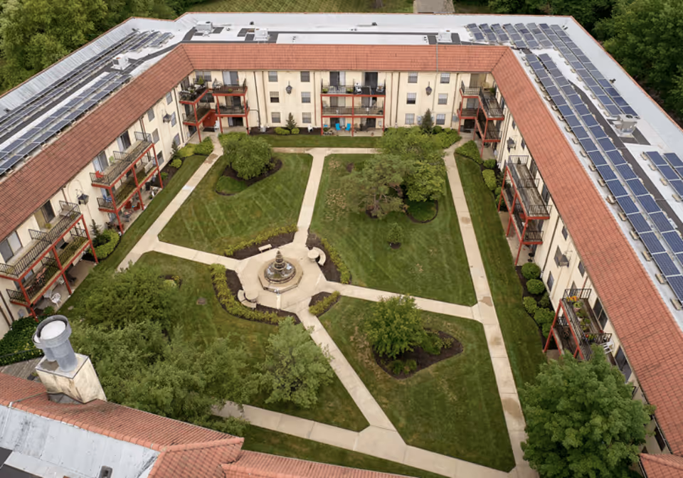 Aerial view of a senior living facility courtyard with green lawns, trees, a central fountain, and surrounding pathways. The building has balconies and solar panels on the roof.