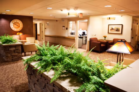 A cozy interior common area with beige walls and carpet, featuring a stone planter filled with green ferns in the foreground. There are comfortable chairs and a table with a lamp, a large wall clock, and framed artwork on the walls. The space is warmly lit with ceiling lights and a table lamp, and an open doorway leads to an office area with a computer.