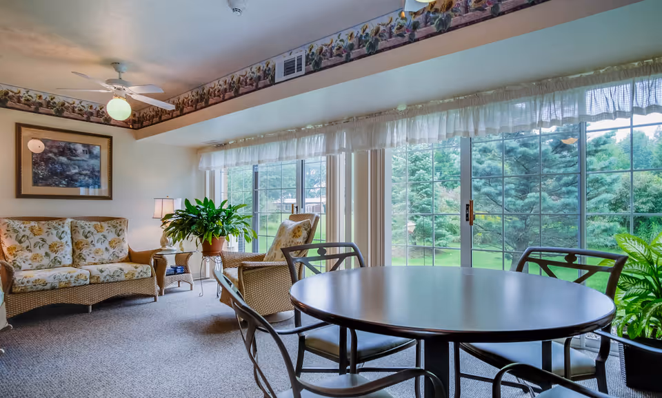 A cozy living room area with floral patterned cushioned wicker sofa and chairs, a round table with metal chairs, large windows with sheer curtains overlooking a green outdoor space with trees, and a ceiling fan with a light fixture.