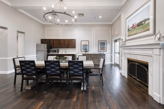 A spacious dining room with a large rectangular table surrounded by eight chairs. The room features dark wood flooring, a modern chandelier, a fireplace with a white mantel, and a large framed picture above it. In the background, there is a kitchenette area with a refrigerator, microwave, and cabinets, along with a sideboard and two framed pictures on the wall.