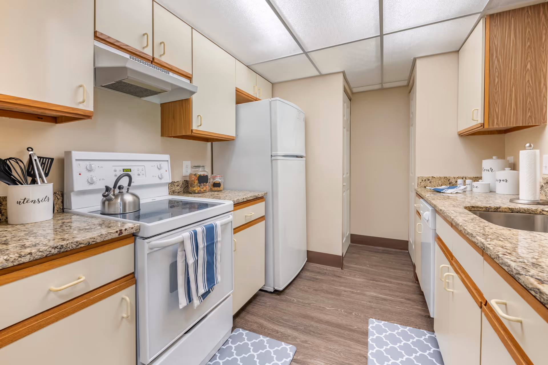 A clean and well-lit kitchen with beige cabinets and granite countertops. The kitchen features a white electric stove with a kettle on it, a white refrigerator, a dishwasher, and a stainless steel sink. There are kitchen utensils in a container labeled 'utensils' on the counter, along with jars and canisters labeled flour, sugar, and tea. Two patterned floor mats are placed on the wooden floor.