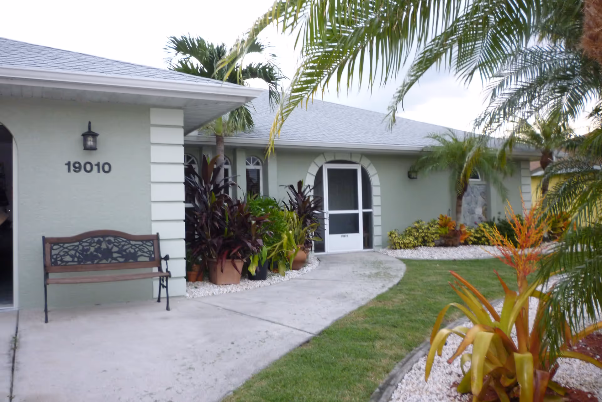 Exterior view of a single-story building with light green walls and a gray roof. The building has the number 19010 displayed near the entrance, which features a bench, potted plants, and a walkway leading to a screened door. Palm trees and other tropical plants surround the area.