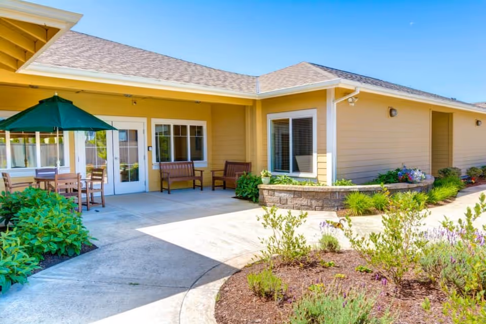 Sunny courtyard patio with seating, a green umbrella, and landscaped flower beds in front of a yellow single-story memory care residence.