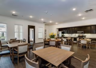 A spacious dining room with multiple wooden tables and chairs arranged neatly. The room has large windows allowing natural light to enter, and a kitchen area with dark cabinetry and stainless steel appliances is visible in the background. The ceiling has recessed lighting, and there is a door and some wall decor on one side.