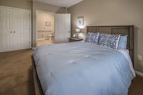 A bedroom with a neatly made bed featuring a light blue comforter and decorative pillows with blue patterns. There is a wooden headboard, a nightstand with a lamp and a clock, and a framed picture on the wall. The room has beige walls and carpeted floor. An open door reveals a bathroom with a toilet and towel holder.