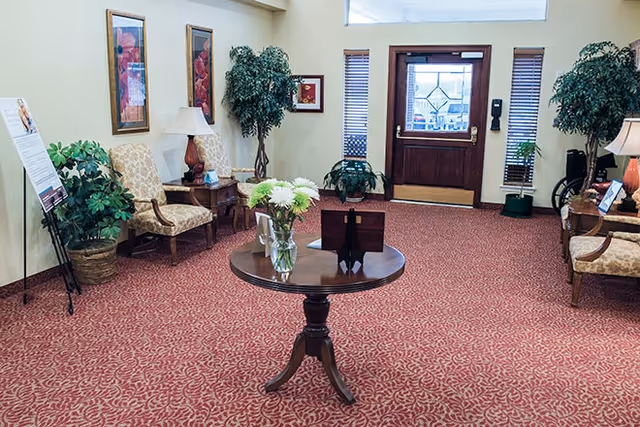 A cozy interior common area with patterned red carpet, a round wooden table with a vase of flowers in the center, two upholstered chairs with a small table and lamp between them, potted plants, framed artwork on the walls, and a wooden door with glass panels at the far end.